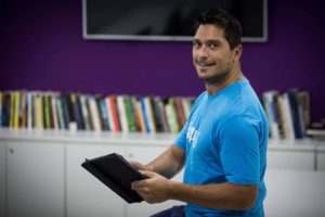 Fundador da startup AltruS, Bruno Aleixo, no escritório da Sevensete de camiseta azul e com o um tablet na mão. Ao fundo uma biblioteca e uma televisão na parede roxa.